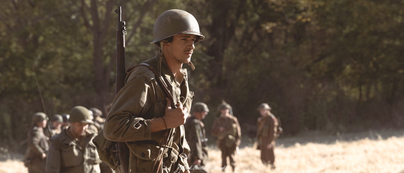 "A portrait of an American soldier at the Lauer Farms WWII reenactment, standing proudly in period-accurate uniform. The soldier's expression is solemn, reflecting the intensity and sacrifice of those who fought in World War II. His gear is detailed, including a helmet and combat pack, with the historical reenactment camp visible in the background. The scene transports viewers back in time, paying homage to the bravery and commitment of U.S. troops."